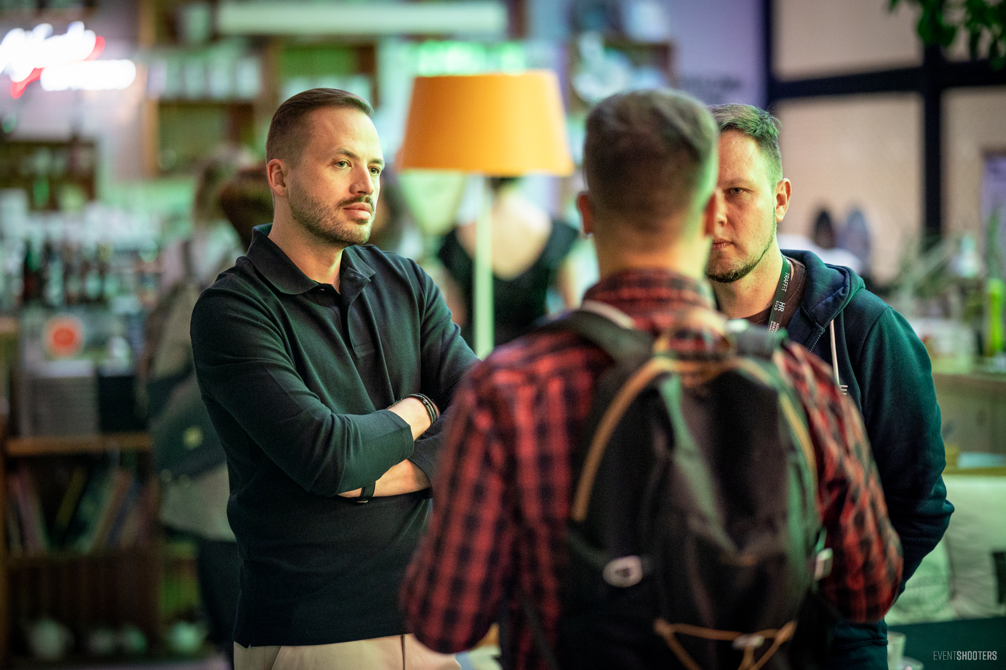 In the photo, three men are having a conversation in a relaxed, networking atmosphere. One of them, wearing a black T-shirt, stands with his arms folded and a focused expression on his face. The second, wearing a hooded sweatshirt with a lanyard around his neck, looks at the interviewer. The third, seen from behind, is wearing a rucksack and a checked shirt.