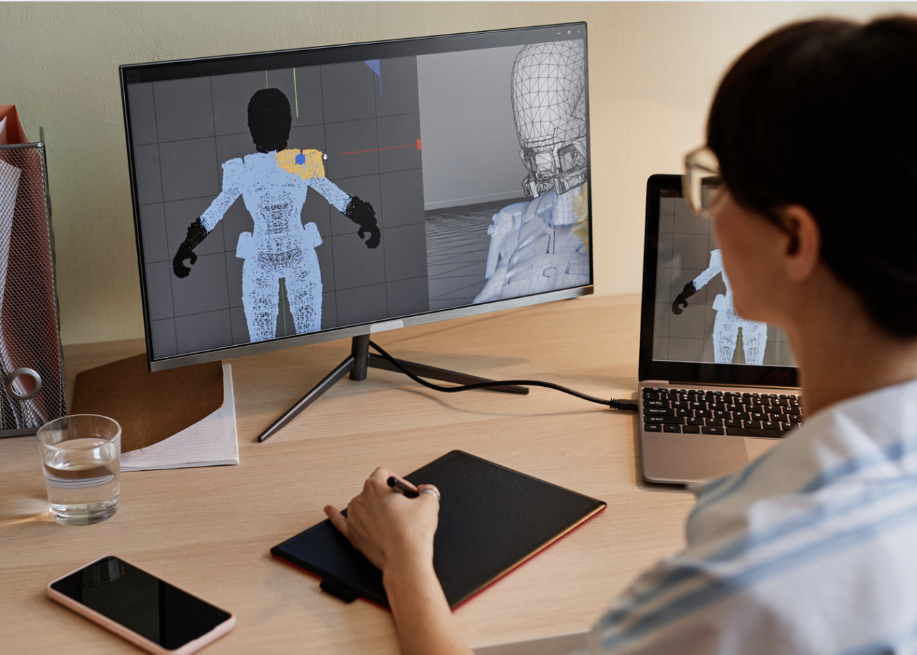 A woman wearing glasses works at a desk using a graphics tablet. On the computer monitor, a 3D character model in a geometric grid is visible and being edited.